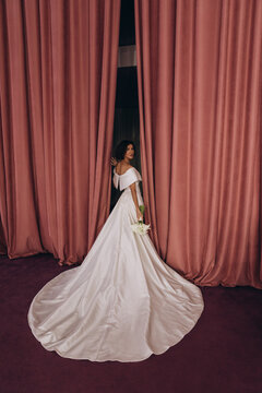 Elegant bride in a long train wedding gown holding flowers while posing gracefully between tall pink velvet curtains. Bride in Flowing Gown Holding Flowers by Velvet Curtains