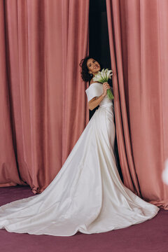Elegant bride in a long train wedding gown holding flowers while posing gracefully between tall pink velvet curtains. Bride in Flowing Gown Holding Flowers by Velvet Curtains