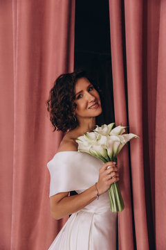 Elegant bride in a long train wedding gown holding flowers while posing gracefully between tall pink velvet curtains. Bride in Flowing Gown Holding Flowers by Velvet Curtains