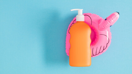 Orange bottle of sunscreen lotion and pink flamingo inflatable toy on blue background. Safe tanning concept on the beach.
