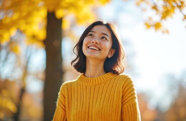 Young Japanese woman with short dark hair smiles, looks up at sunny autumn park. Yellow knitted sweater. Happy female enjoys nature, relaxation. Portrait of joyful person.