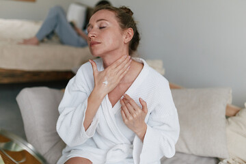 middle aged woman applying lotion on neck in white bathrobe, promoting healthy skin, wellness, and active wellbeing at home during self-care routine
