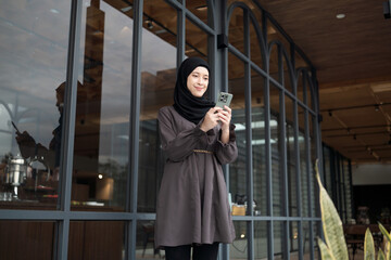A woman wearing a black headscarf and smiling while using her smartphone in front of a modern glass...