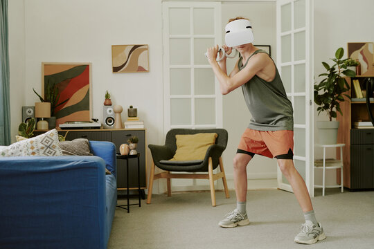 Caucasian teenage boy wearing VR headset standing in living room holding game controller simulating sports activity engaging in virtual reality gaming during leisure time