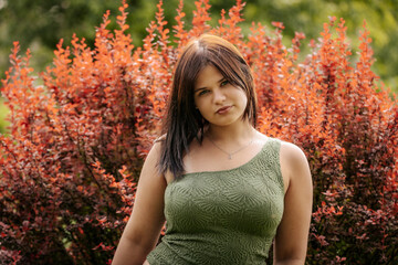 Smiling young woman in a textured green one-shoulder top poses in front of vibrant red foliage,...