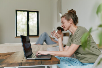 Middle age woman drinking water and using laptop in cozy home environment, promoting active and...