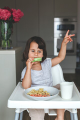 Little girl sitting in high chair eating cucumber and pasta, showing peace sign with hand, concept of childhood and healthy eating, vertical
