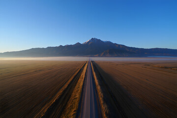 Fototapeta premium Aerial View: Serene Country Road Leading to Majestic Snow-Capped Mountains at Sunrise