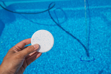 Hand holding a chlorine tablet above a clear blue swimming pool during routine maintenance under...