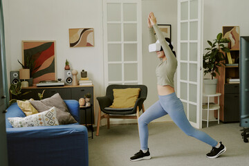Teenage Caucasian girl wearing VR headset exercising in living room, performing yoga pose with arms...
