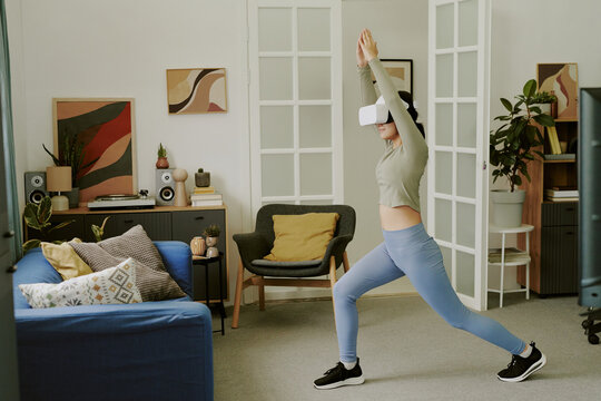 Teenage Caucasian girl wearing VR headset exercising in living room, performing yoga pose with arms raised, engaging in virtual reality fitness activity at home