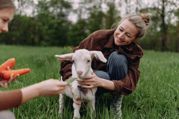 Smiling middle age woman caring for a young goat outdoors, promoting active and healthy lifestyle, wellbeing, community, and wellness in a natural green environment.