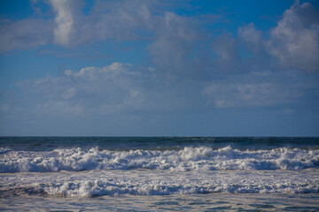 A dynamic seascape captures foamy waves breaking on a sandy shore under a brilliant blue sky dotted...