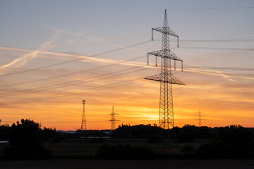 An electricity pylon stands tall as power lines stretch across a vibrant evening sky, with pink and orange hues illuminating layered clouds above a leafy horizon