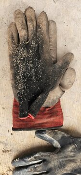 Close-up of worn work gloves with red cuffs and black fabric, covered in dust and lying on a rough wooden surface with visible grain and texture; work gloves.
