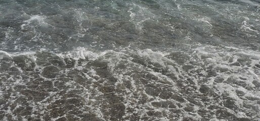 Gentle ocean waves create white foam over clear shallow water, exposing the sandy seabed beneath soft gray and blue hues in natural light; ocean, shoreline.