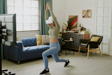 Teenage Caucasian girl exercising with VR headset in living room, performing lunge in front of...