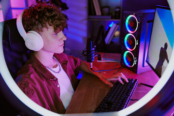 Caucasian teenage boy wearing headphones sitting at desk live streaming video games using desktop computer with RGB lighting and microphone visible in home environment