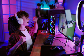 Caucasian teenage boy wearing headphones sitting at desk live streaming video game, smiling and raising fist in excitement, gaming computer and microphone visible in home environment