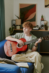 Caucasian teenage boy sitting on sofa playing acoustic guitar at home, looking away from camera,...