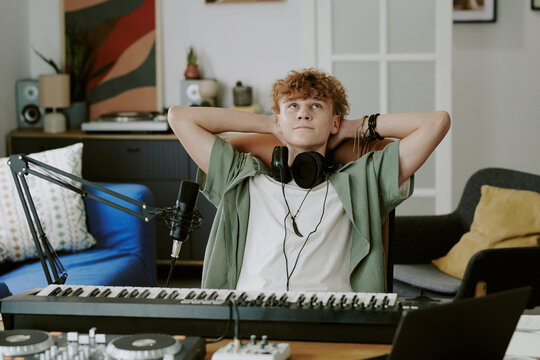 Caucasian teenage boy relaxing with hands behind head sitting at digital piano wearing headphones in home studio recording music surrounded by audio equipment