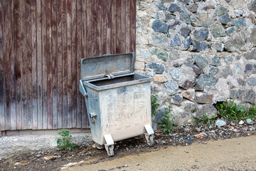 Old dirty metal trash bin with open lid standing on a muddy street near stone and wooden wall,...