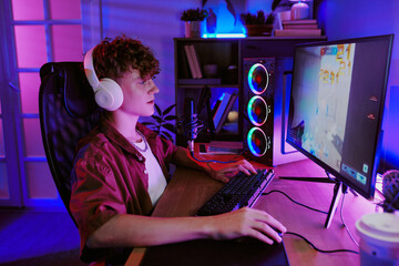 Caucasian teenage boy wearing headphones sitting at desk live streaming video games on desktop computer with microphone visible, focused on gaming in modern home environment