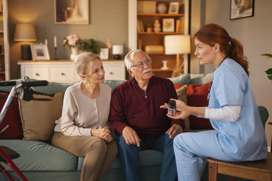 In a cozy living room, a friendly nurse in blue scrubs sits and shows a pill bottle to a senior couple on a sofa. The sunlight streams through the window, creating a warm, supportive atmosphere.