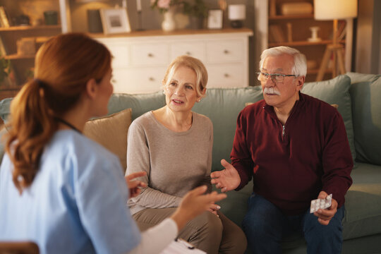 A female home healthcare nurse, dressed in scrubs, is explaining medication to a senior couple. The couple, seated on their couch, listens attentively during a daytime visit in their Living Room.