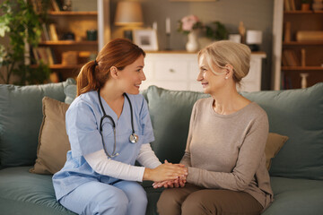 A young female home nurse wearing scrubs and stethoscope sits on a sofa with an elderly woman and...