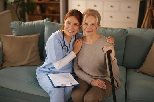 A home healthcare nurse in scrubs smiles while embracing a senior woman who is holding a cane. They are sitting on a sofa in the woman's living room during a daytime visit.