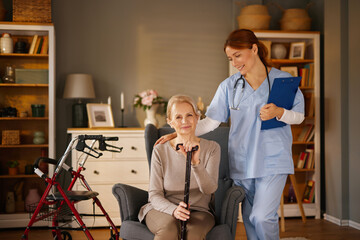 A female nurse, wearing a blue uniform and stethoscope, smiles as she places her hand on the...