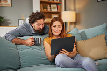 Relaxed couple spending quality time together in their cozy living room. They are seated on a comfortable couch, with one partner sipping coffee while looking at a tablet.