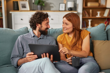 A couple sits closely together on a couch, sharing smiles and laughs as they look at a tablet. The...