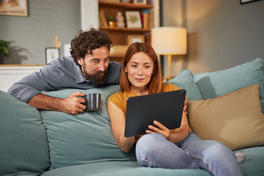 Relaxed couple spending quality time together in their cozy living room. They are seated on a comfortable couch, with one partner sipping coffee while looking at a tablet. - Powered by Adobe