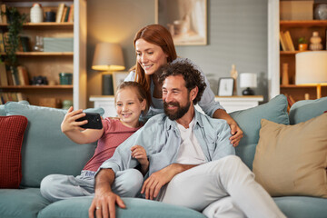 A family of three smiles as they pose for a selfie, sitting comfortably on a couch surrounded by a...