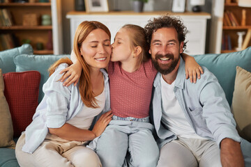 A happy family enjoys a loving moment on the couch. The child kisses one parent while the other...