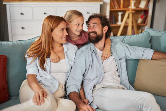 A happy family sits closely together on a couch in a warm living room. The parents share smiles with their daughter, creating a joyful atmosphere filled with love and affection.