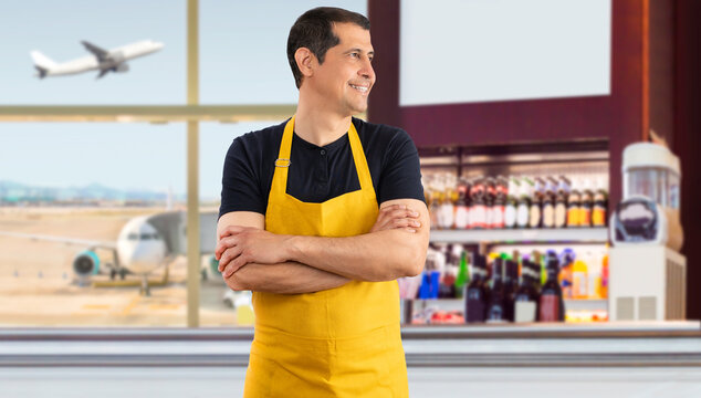 Hispanic barista standing smiling looking to the side and staring away thinking with yellow apron in the coffee shop at an airport
