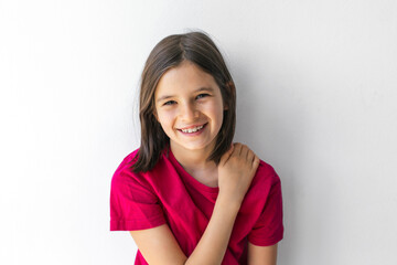Cheerful child girl 8-9 years with big smile and hand on shoulder against white wall, concept of childhood happiness and self-confidence