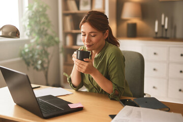 A woman sits at her home office desk, savoring a warm drink. Morning light fills the room,...