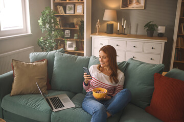 A woman relaxes on a comfortable couch, eating a bowl of snacks while looking at her phone and laptop in a warm living room filled with natural light.