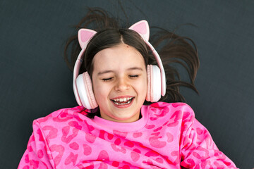 Smiling girl lying down with eyes closed, wearing pink headphones with cat ears and pink leopard sweatshirt, enjoying music, concept of childhood and joy