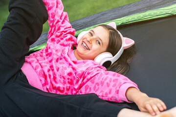 Cheerful child girl 8-9 years laughing on trampoline in pink cat ear headphones and sweatshirt, concept of joy and childhood play