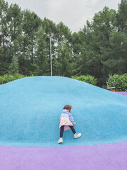 Little girl climbing a bright blue rubber hill at colorful modern playground surrounded by green trees, concept of childhood adventure and active outdoor play