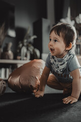 A joyful and curious kid in a gray shirt and beige shorts, smiling and looking up, in a home environment next to a copper balloon