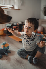 A joyful toddler sitting on a wooden floor, reaching towards the dog, in a bright, homely setting full of toys