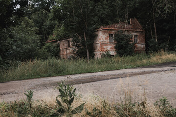 An abandoned, old brick house overgrown with nature, standing on a dirt road, with a blurred background