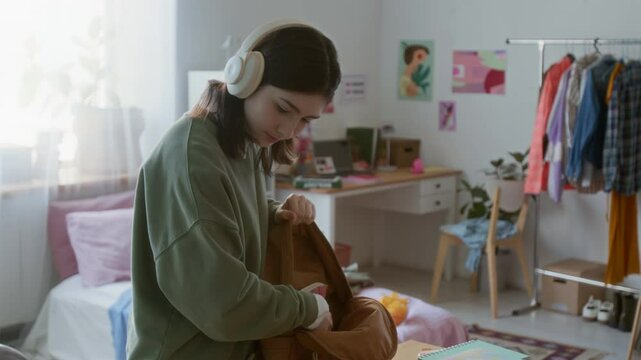 Pan up shot of Caucasian teenage girl packing backpack in dorm room and wearing big headphones