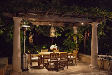Obraz premium Charming outdoor space, dining table, pergola, statue, columns, beautifully lit at night. Surrounding greenery enhances the serene atmosphere, cozy and inviting setting for gatherings. 